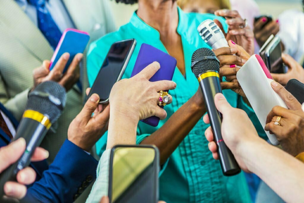 The mid-section of an African-American woman dressed in teal as media phones and microphones are pointed at her.