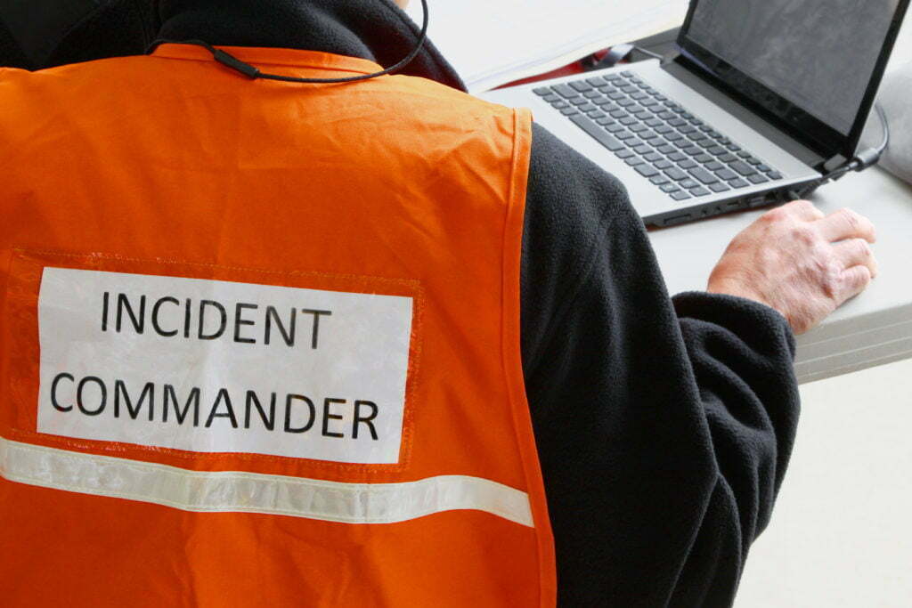 The words "incident commander" adorn the back of an orange vest on an incident commander sitting at a desk, working on a laptop computer.