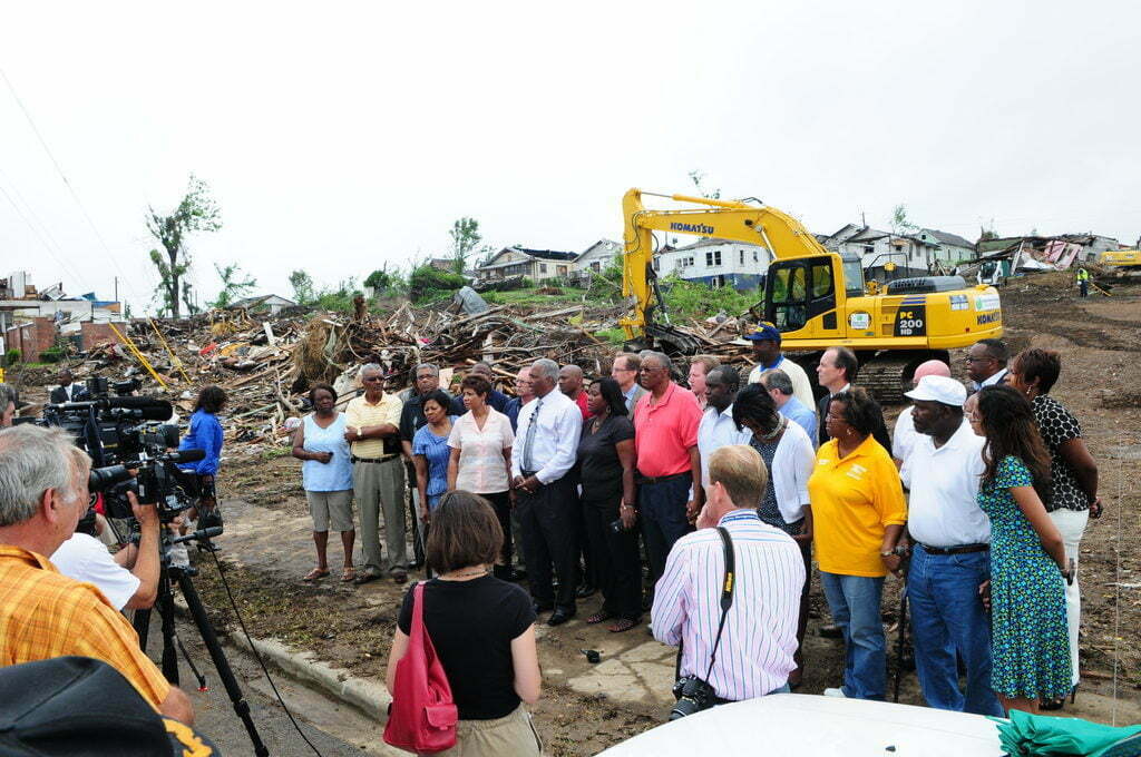 City officials stand in front of disaster debris and heavy machinery speak to media about storm recovery.