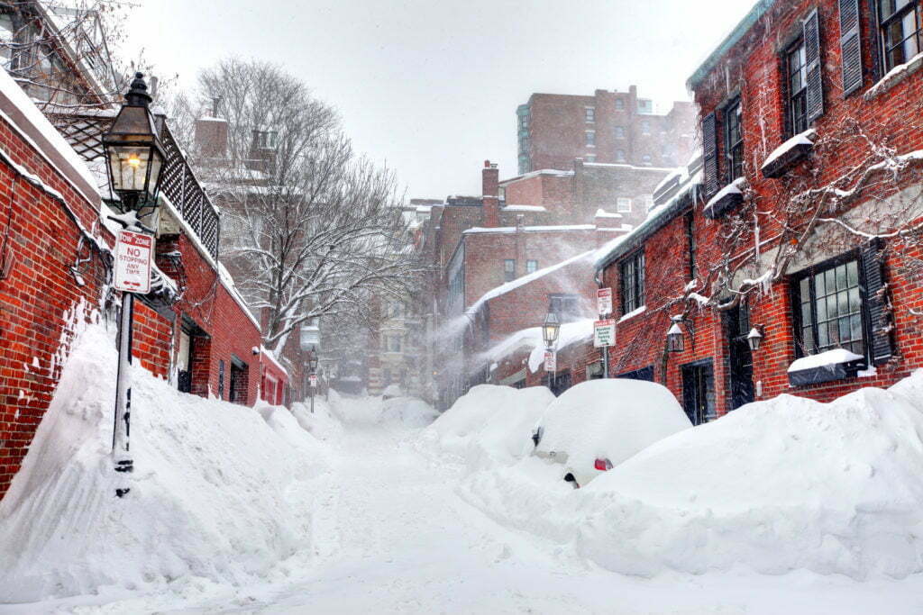 A view down a brick-apartment lined street with snow blowing after a snowstorm and steep snowbanks on both sides of the street..
