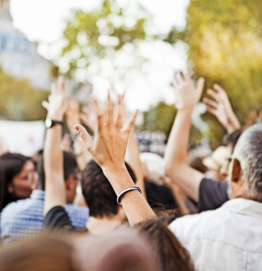 A crowd gathered outside at a press conference that is raising hands to ask a question to the speaker.