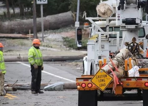 Utility workers oversee a utility truck loading downed utility poles for removal from a street littered with downed power lines and debris.