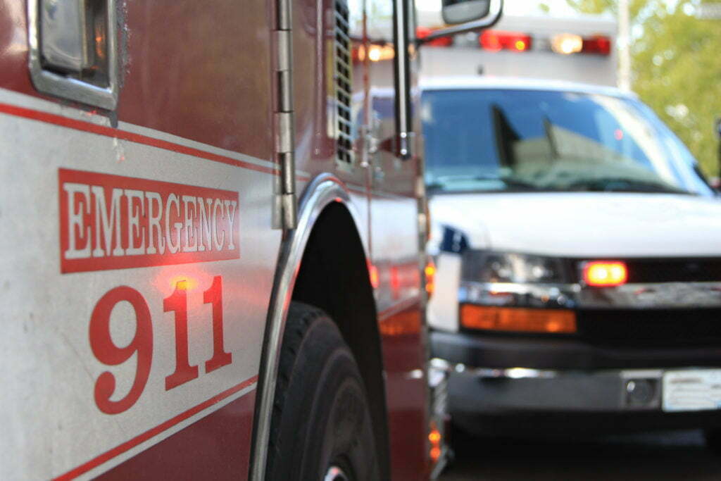 An emergency scene showing a closeup of the text "Emergency 911" on a firetruck with an ambulance in the background.
