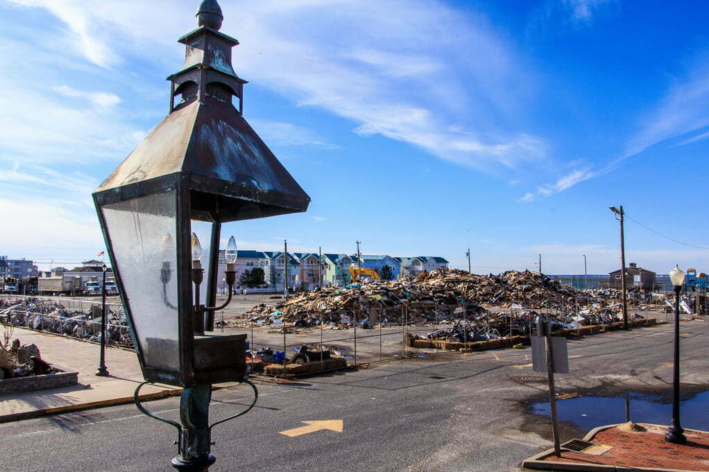 A broken lamppost in the forefront; piles of debris sit in a fenced parking lot in the background.