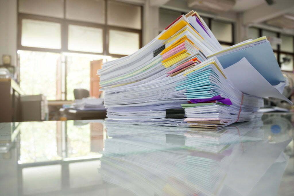 Pile of unfinished documents on office desk, held together with clips and rubber bands.