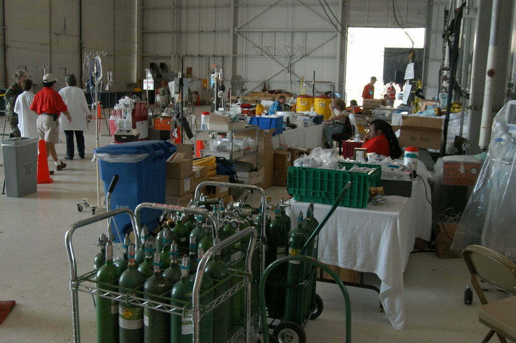 Piles of medical supplies sit in a warehouse as doctors and volunteers sit at tables and walk about.